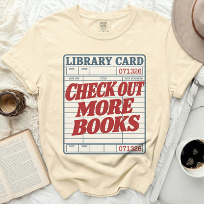 Light yellow t-shirt with a library card graphic reading "Check Out More Books."