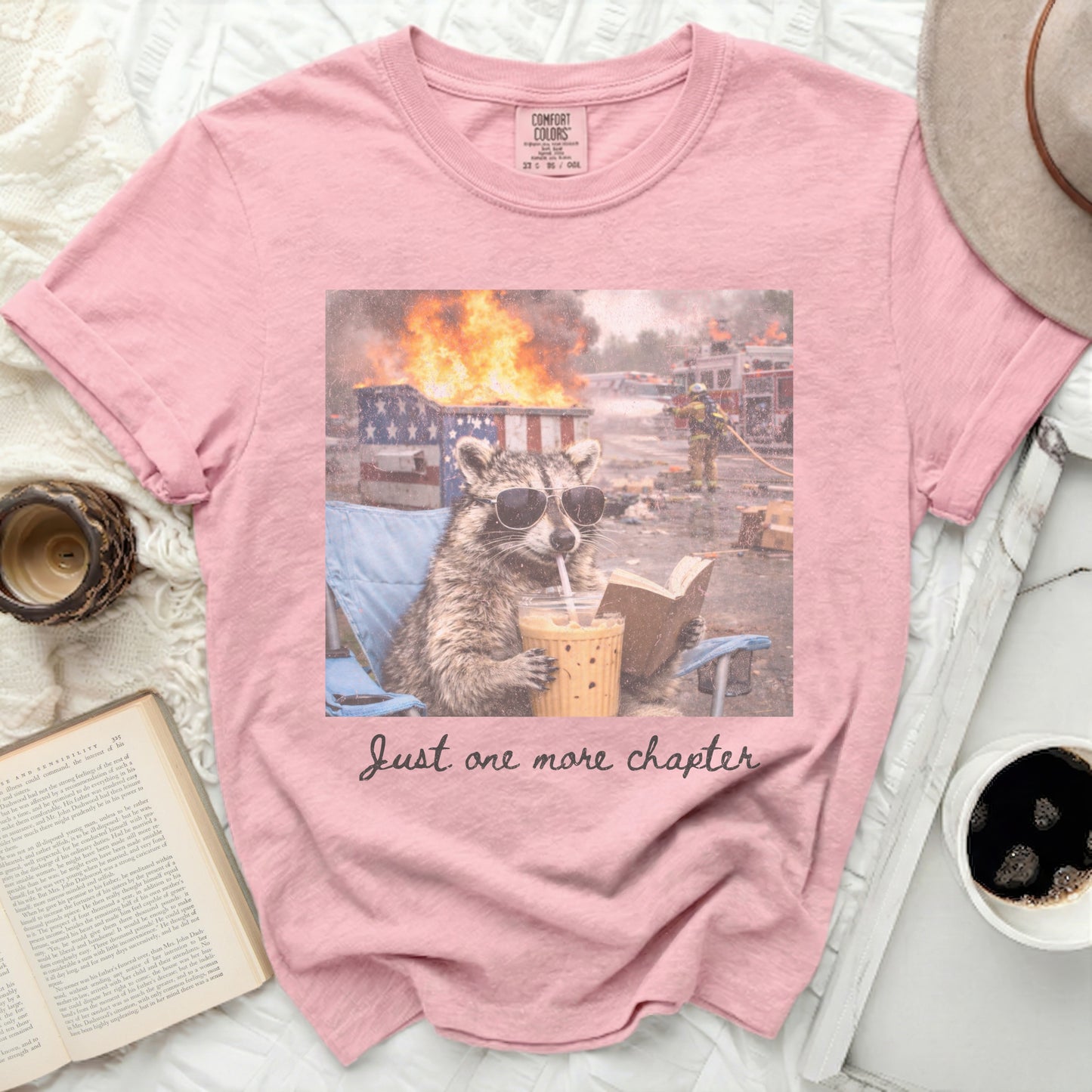 Pink t-shirt with sunglasses-wearing raccoon holding iced coffee, fires in background.