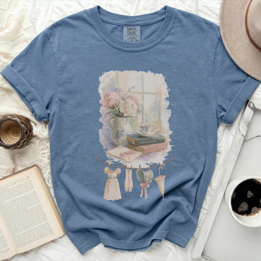 Blue t-shirt with watercolor print of flowers, books, and vintage clothing on a line.