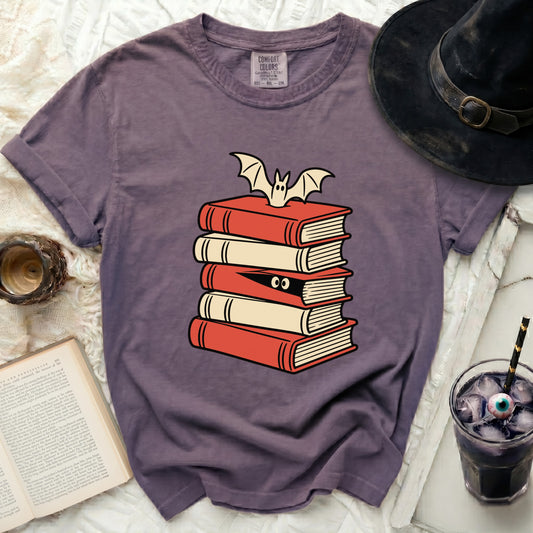 Purple t-shirt with stacked books, bat, and peeking eyes graphic.