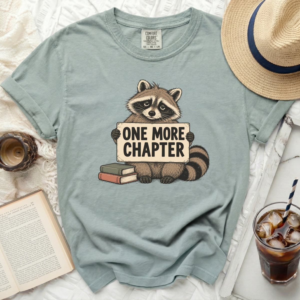 Light blue t-shirt with raccoon holding "ONE MORE CHAPTER" sign.