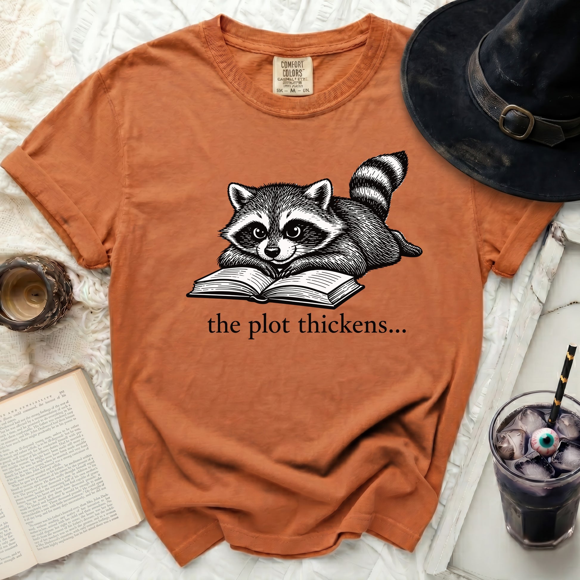 Orange t-shirt with a raccoon reading a book, text "the plot thickens..."