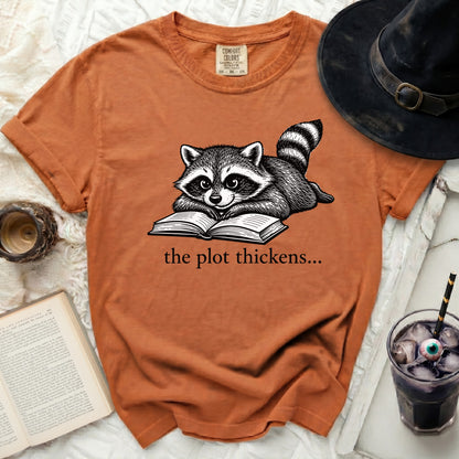 Orange t-shirt with a raccoon reading a book, text "the plot thickens..."