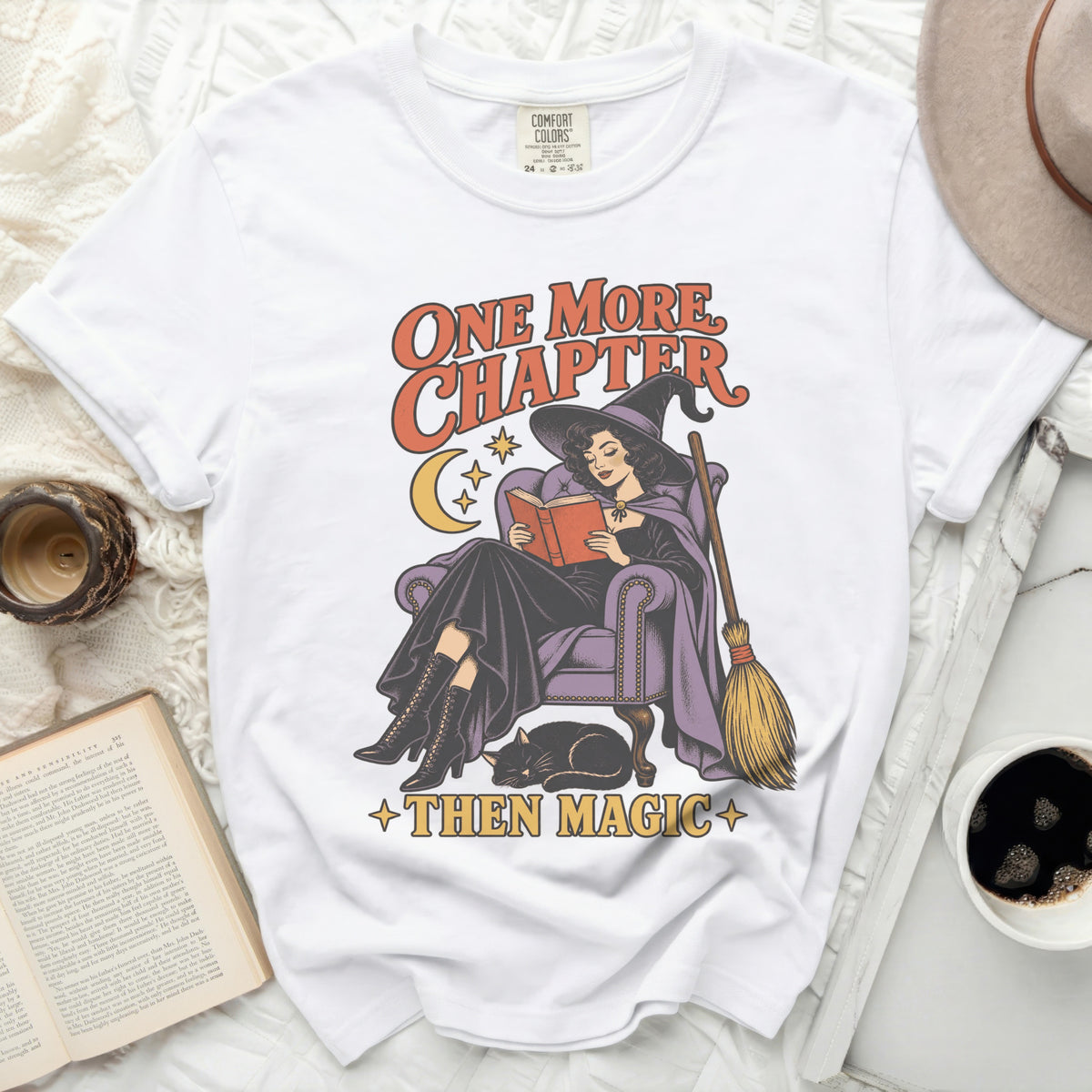 White t-shirt with graphic of witch reading, broom, cat, and text "One More Chapter Then Magic".