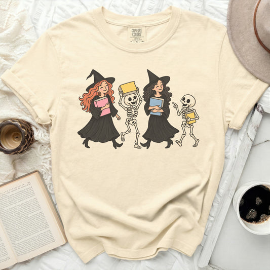 Two witches and a skeleton holding books, illustrated on a cream t-shirt.