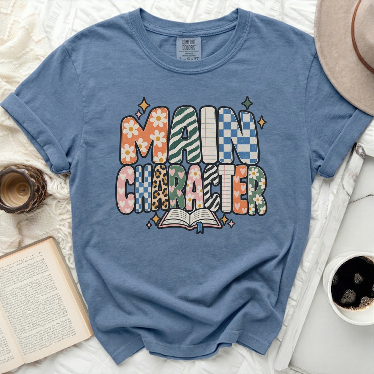 Blue t-shirt with "MAIN CHARACTER" in colorful, patterned letters and an open book.