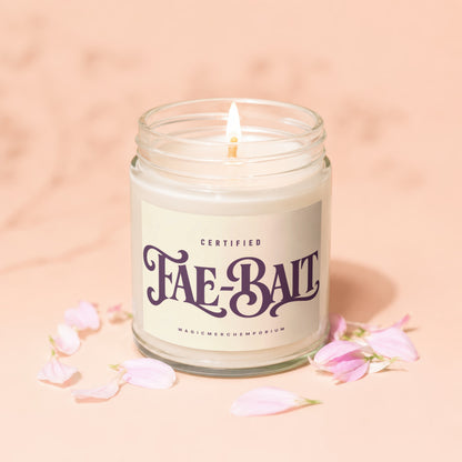 Candle with "Fae-Bait" label, lit wick, and pink petals on peachy background.
