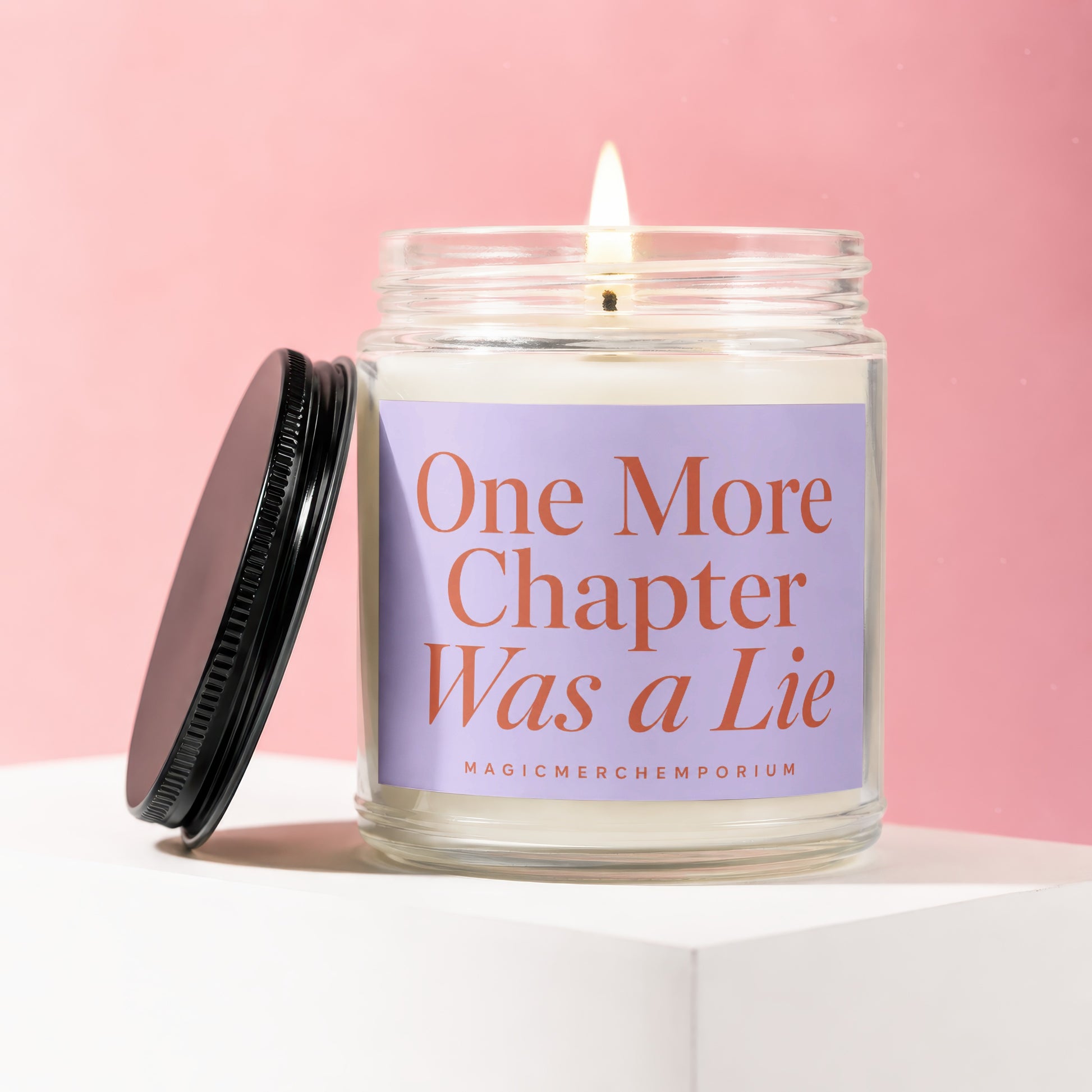 Glass candle with lit wick, black lid, and purple label that reads "One More Chapter Was a Lie."