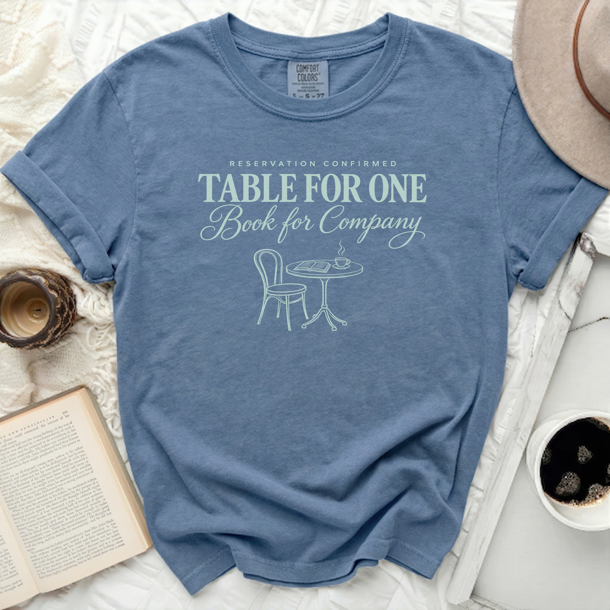 Blue t-shirt with "Table for One Book for Company" graphic and cafe scene.
