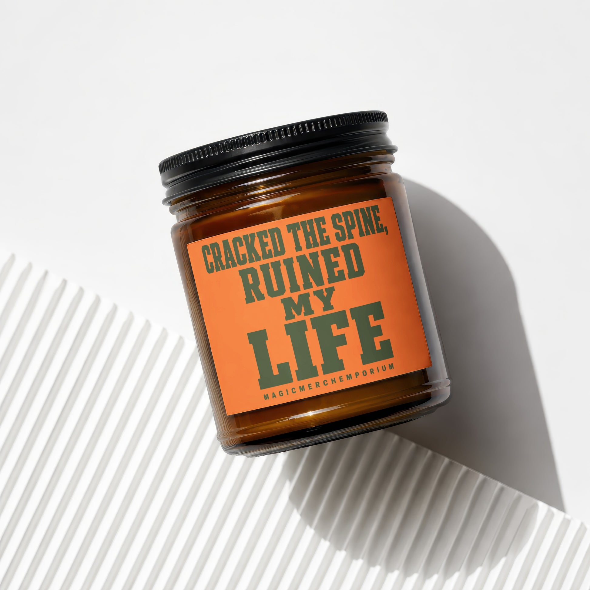 Amber jar candle with orange label reading "CRACKED THE SPINE, RUINED MY LIFE"