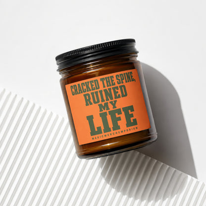 Amber jar candle with orange label reading "CRACKED THE SPINE, RUINED MY LIFE"