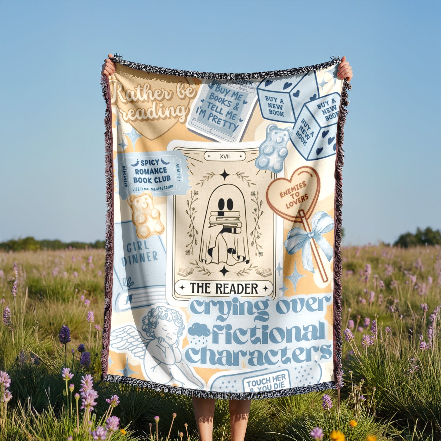A person is holding a colorful blanket with various text and illustrations, including a tarot card, flowers, and a teddy bear, in a field with purple flowers.