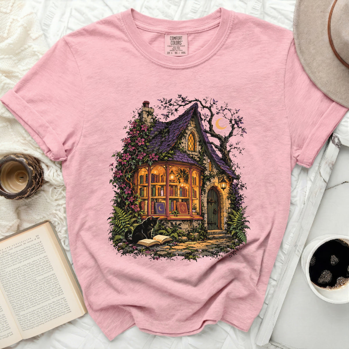 Pink t-shirt with an illustration of a magical cottage, black cat, and open book.