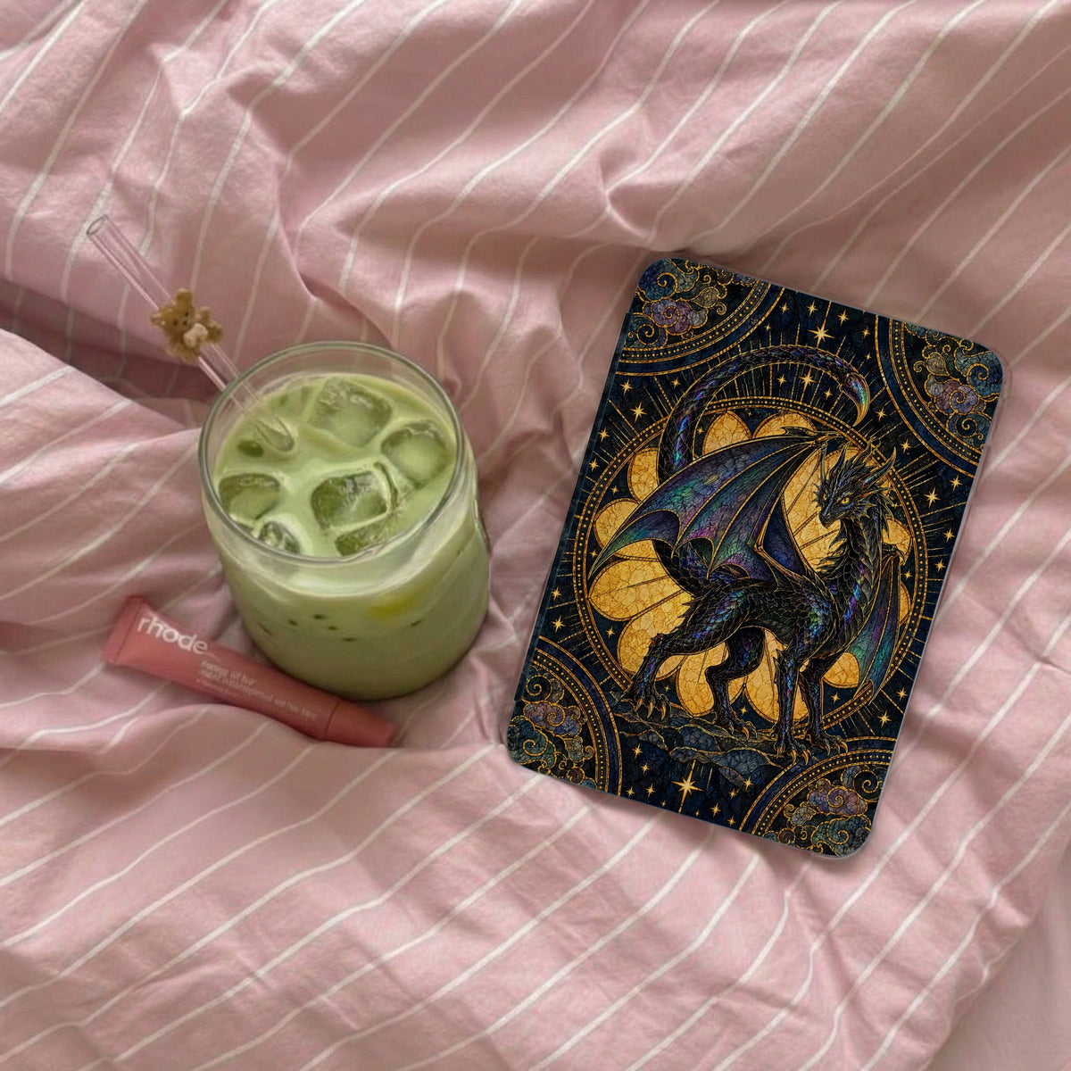 An e-reader with an ornate dragon case, an iced matcha latte, and a pink lip balm rest on pink striped bedding.