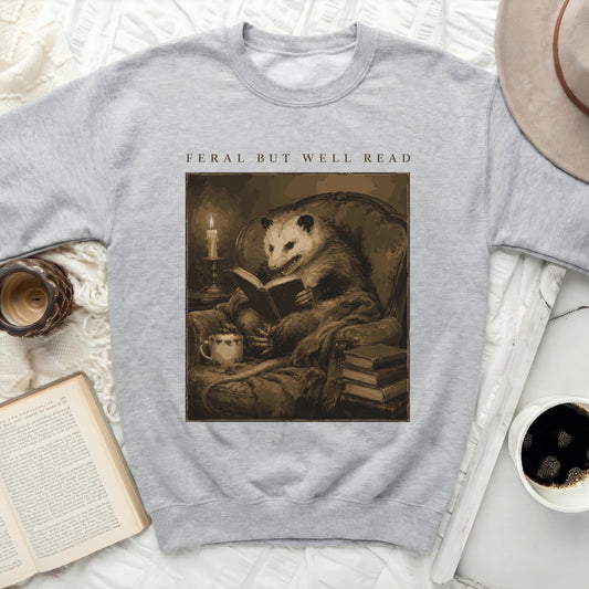 Gray sweatshirt with print of an opossum reading a book titled "Feral but Well Read".