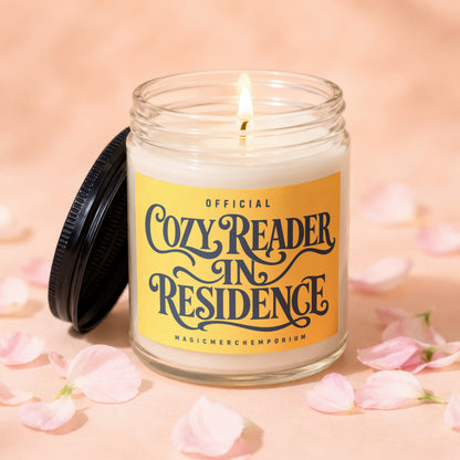 Glass candle labeled "Official Cozy Reader In Residence" with burning wick.