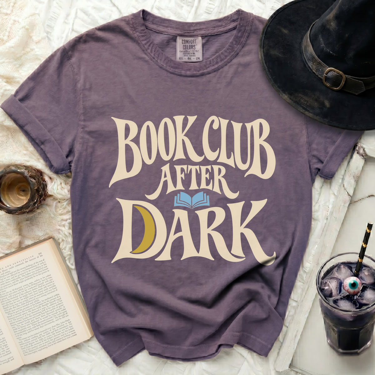 A purple crewneck t-shirt featuring the white text "BOOK CLUB AFTER DARK" with a blue open book and yellow crescent moon graphic.