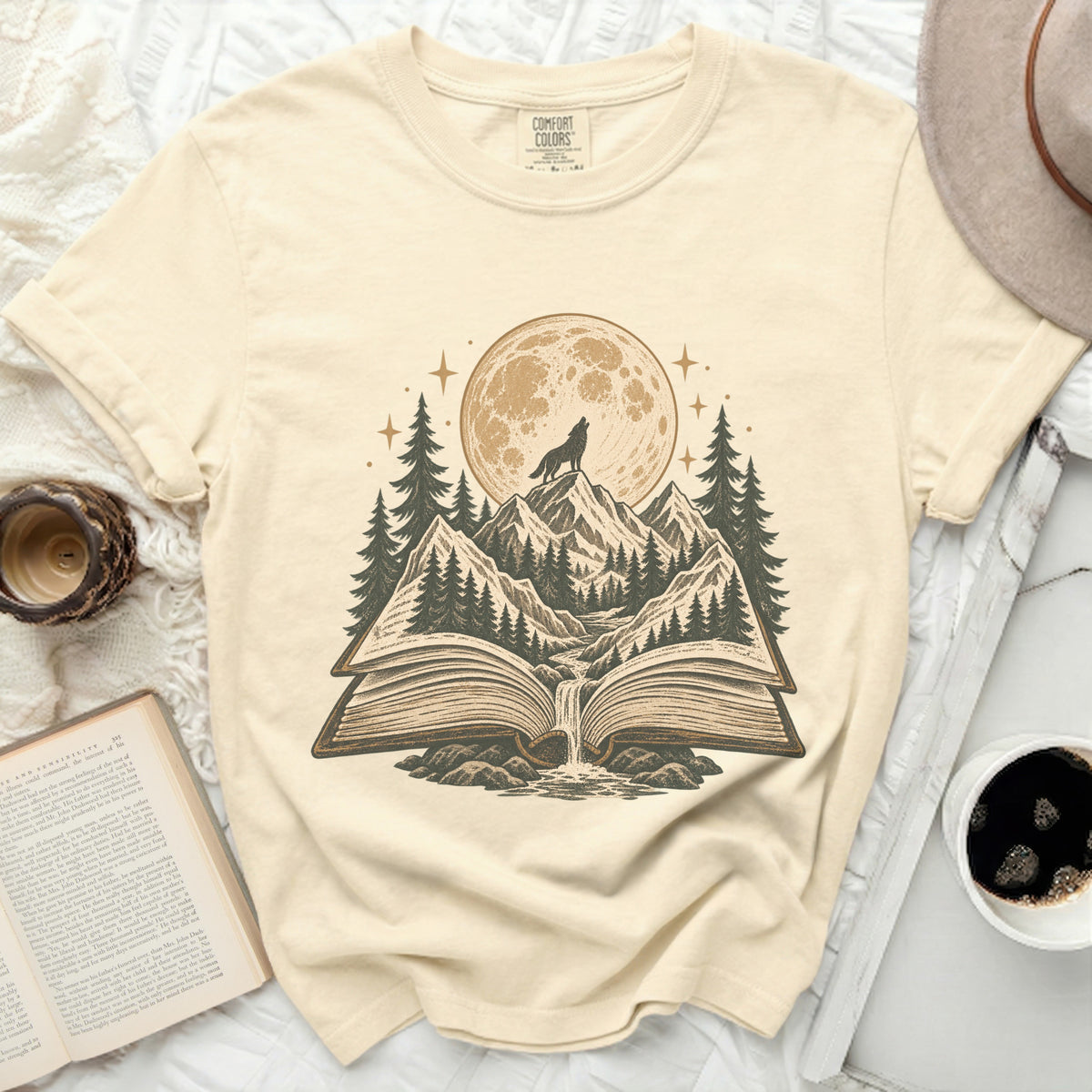 Cream t-shirt with graphic of wolf howling at moon over mountains from an open book.