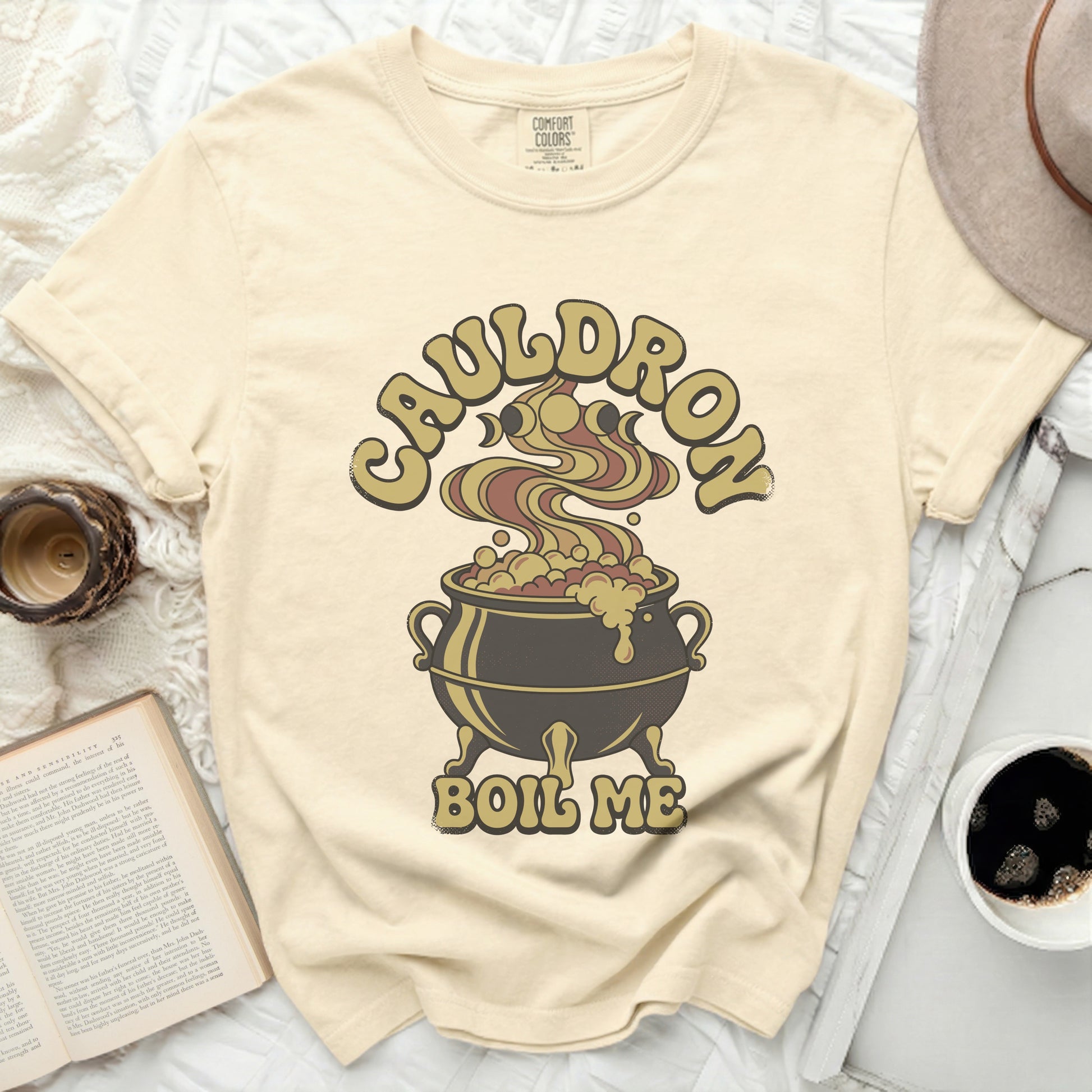 Cream t-shirt with "Cauldron Boil Me" graphic of a cauldron and moon phases.