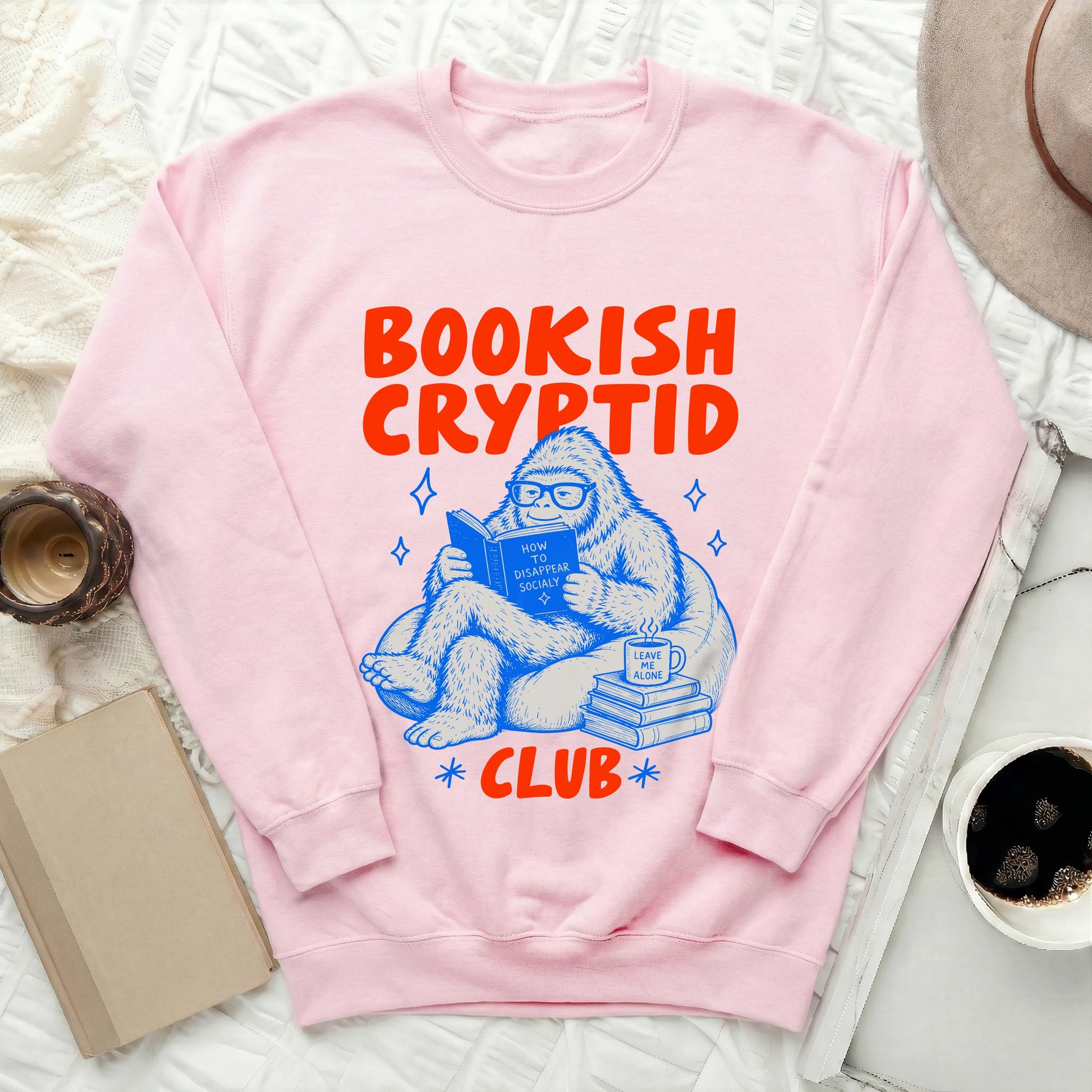 Pink sweatshirt with "Bookish Cryptid Club" text and a Bigfoot reading a book.