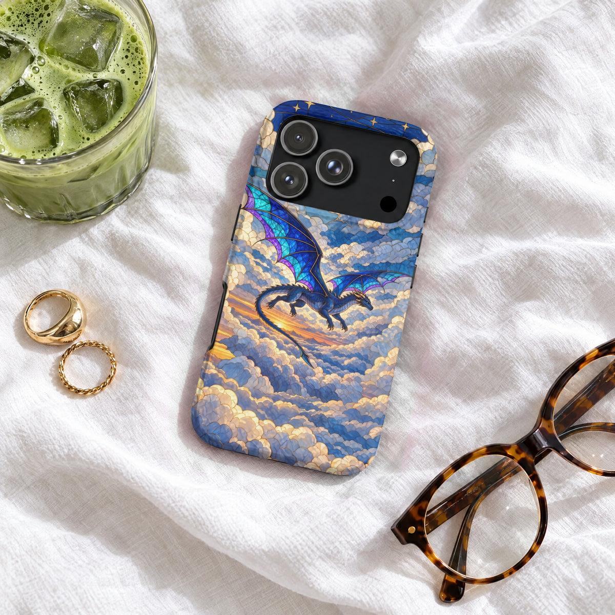 Smartphone case featuring a blue dragon flying through stained glass style clouds on a white surface with glasses and iced matcha.