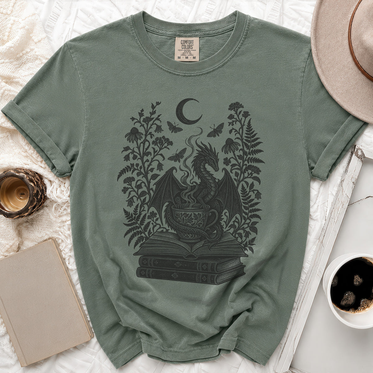 A sage green t-shirt with a black line art graphic of a dragon, teacup, stacked books, and flowers.