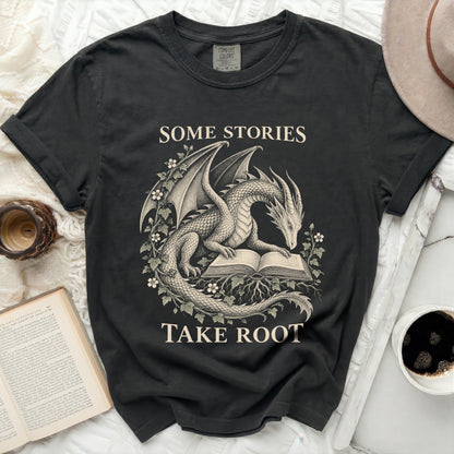 Dark t-shirt with dragon reading a book design and text "Some stories take root".