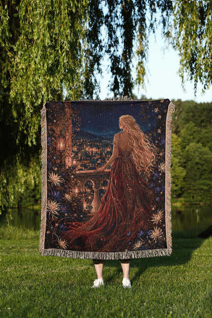 A person stands in a grassy field, holding a large tapestry depicting a woman with long, flowing hair and a flowing red dress, gazing out at a cityscape at night.