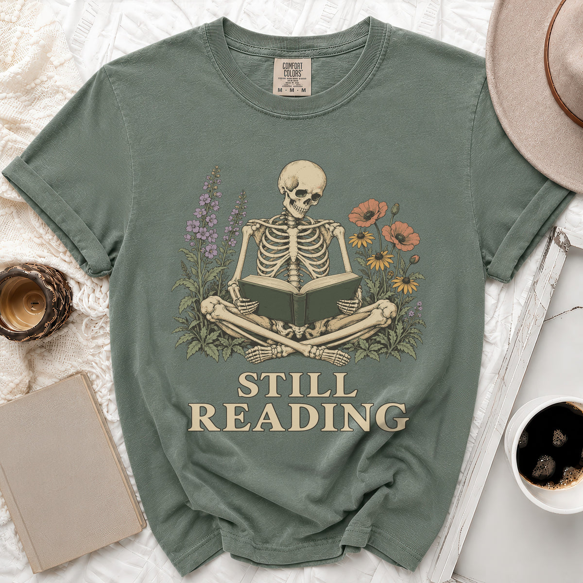 Green t-shirt with skeleton reading a book and floral accents, text "STILL READING".