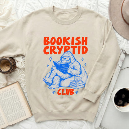 Cream sweatshirt with "Bookish Cryptid Club" text and cartoon Bigfoot reading.
