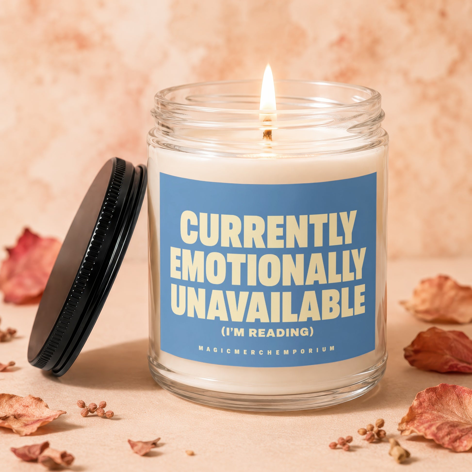 Glass candle with lit wick, blue label reading "CURRENTLY EMOTIONALLY UNAVAILABLE (I'M READING)".