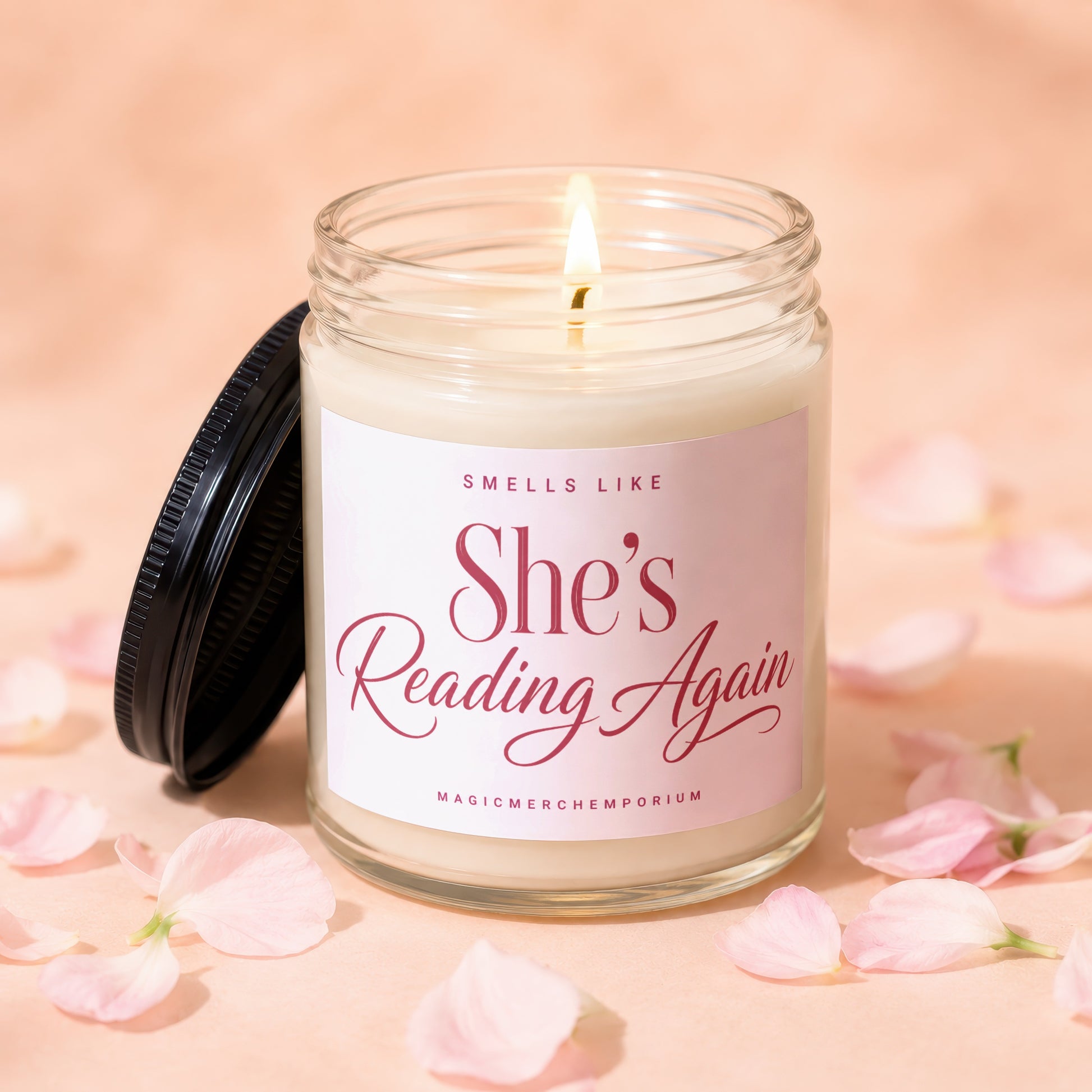 Candle with lit wick, a black lid, and a label that reads "She's Reading Again".