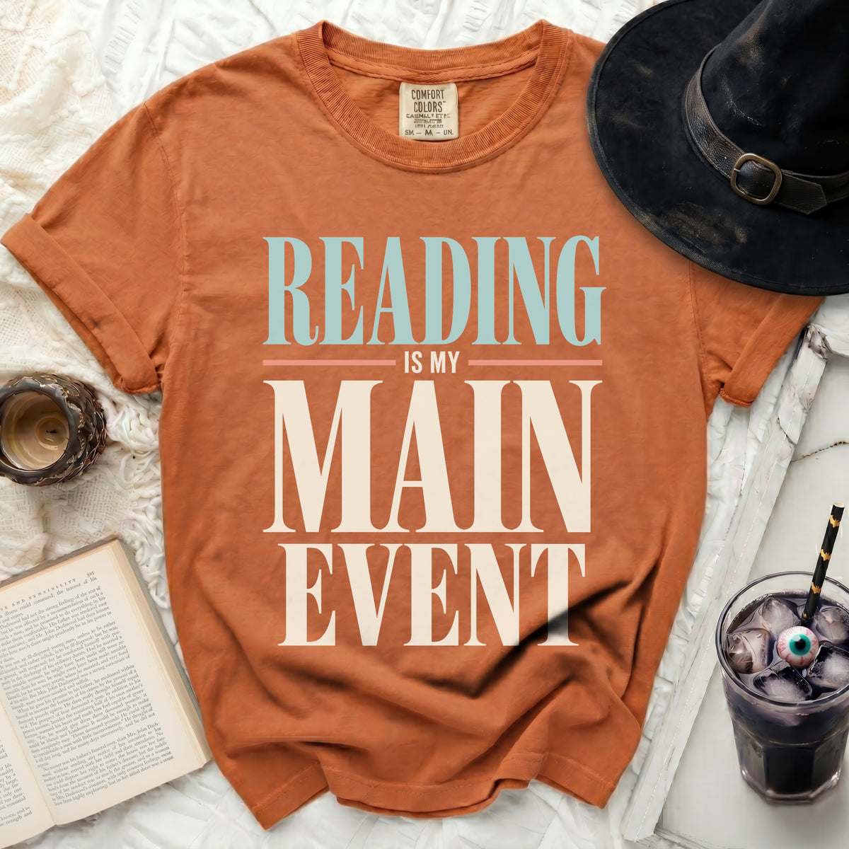 Orange t-shirt with "READING IS MY MAIN EVENT" text, witch hat, book, and drink.