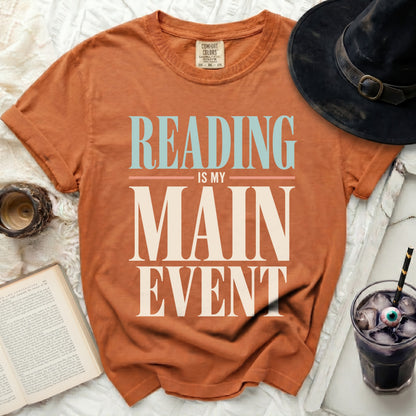 Orange t-shirt with "READING IS MY MAIN EVENT" text, witch hat, book, and drink.
