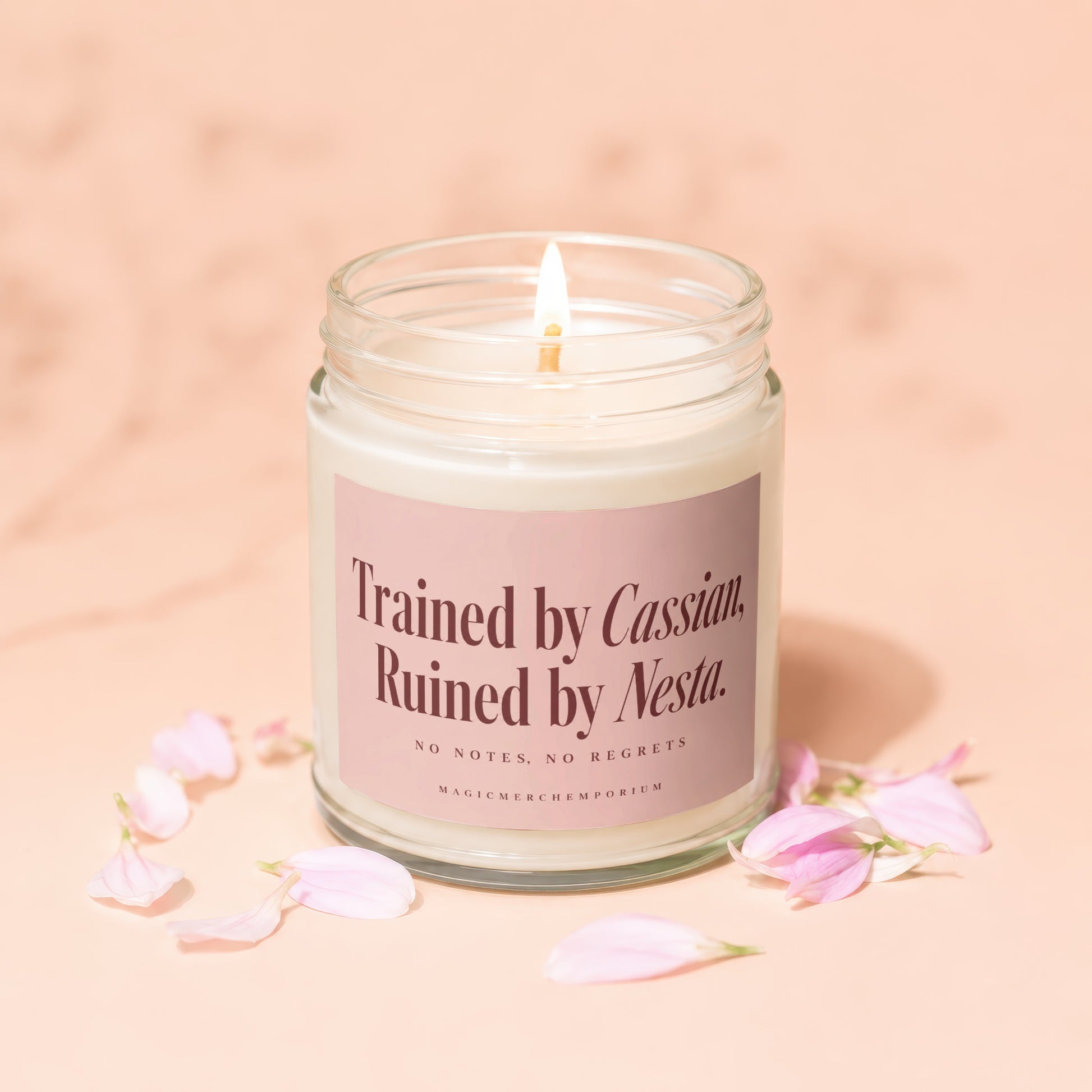 Lit scented candle in a glass jar featuring a pink label with text, surrounded by pink petals.