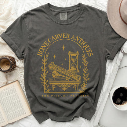 A dark gray crewneck t-shirt featuring a gold print with an hourglass, key, bone, and text reading "Bone Carver Antiques The Prison Prythian."