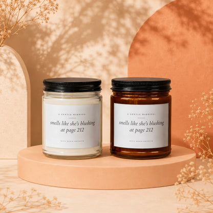 Two candles, one clear and one amber jar, with labels reading "smells like she's blushing at page 212".