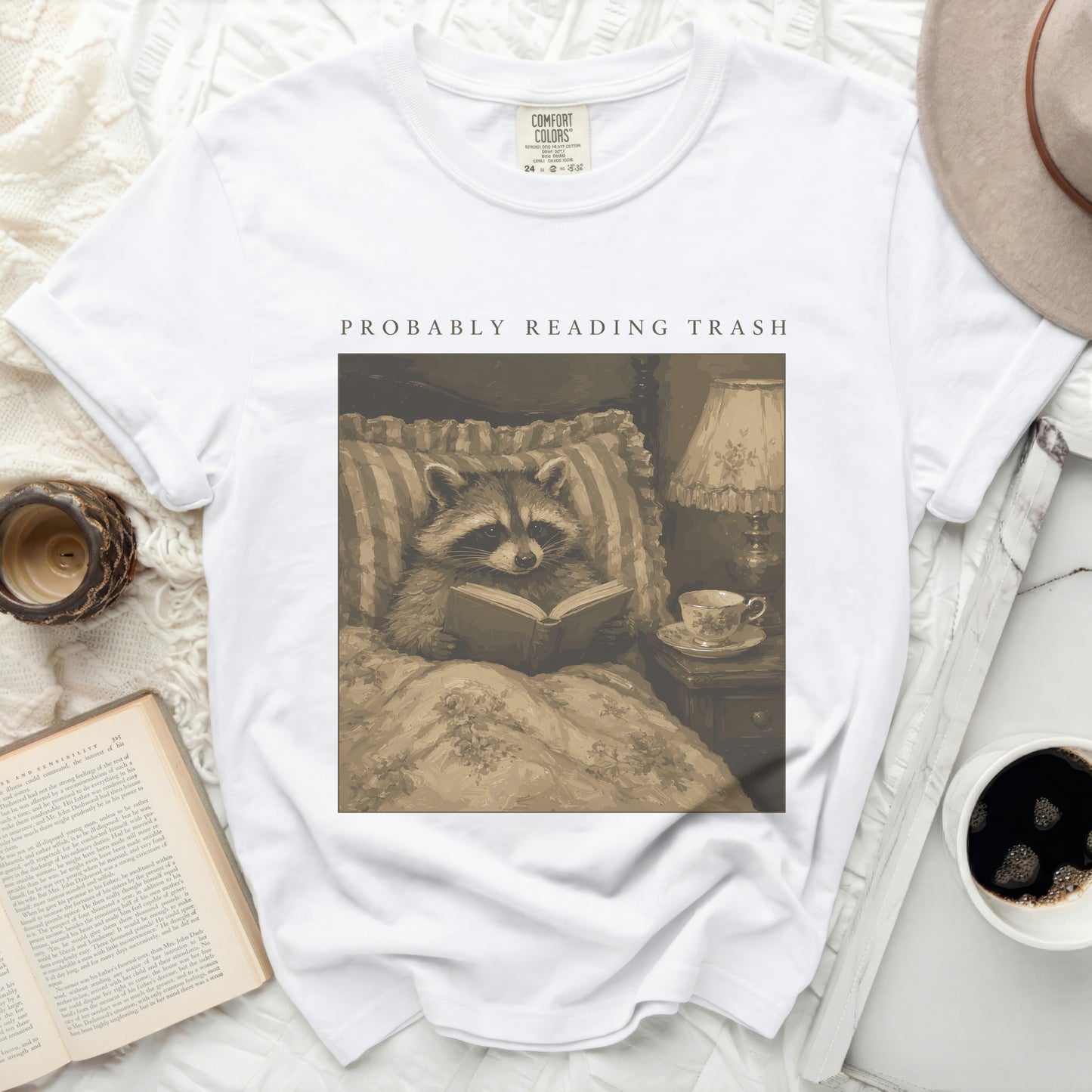 White t-shirt with a print of a raccoon reading in bed with the text "PROBABLY READING TRASH".