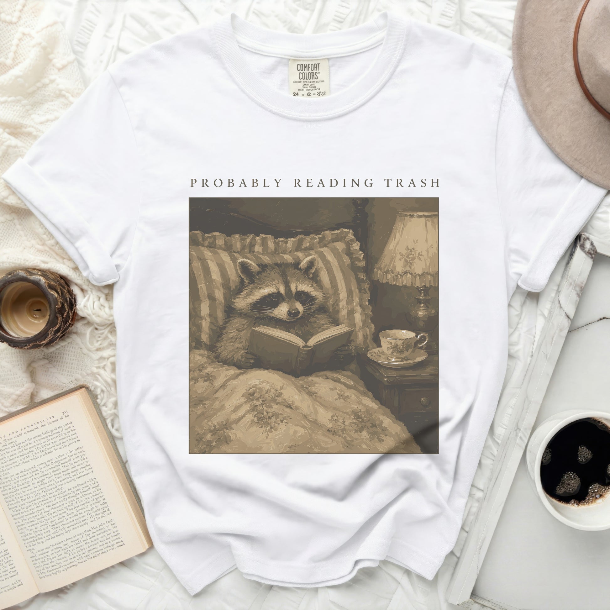 White t-shirt with a print of a raccoon reading in bed with the text "PROBABLY READING TRASH".