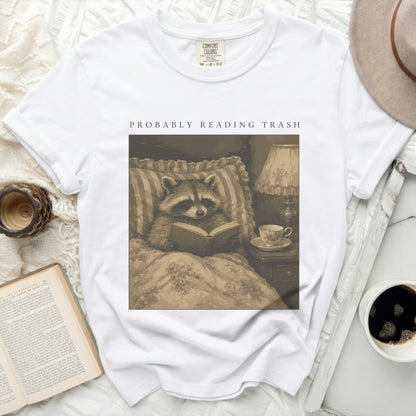 White t-shirt with a print of a raccoon reading in bed with the text "PROBABLY READING TRASH".