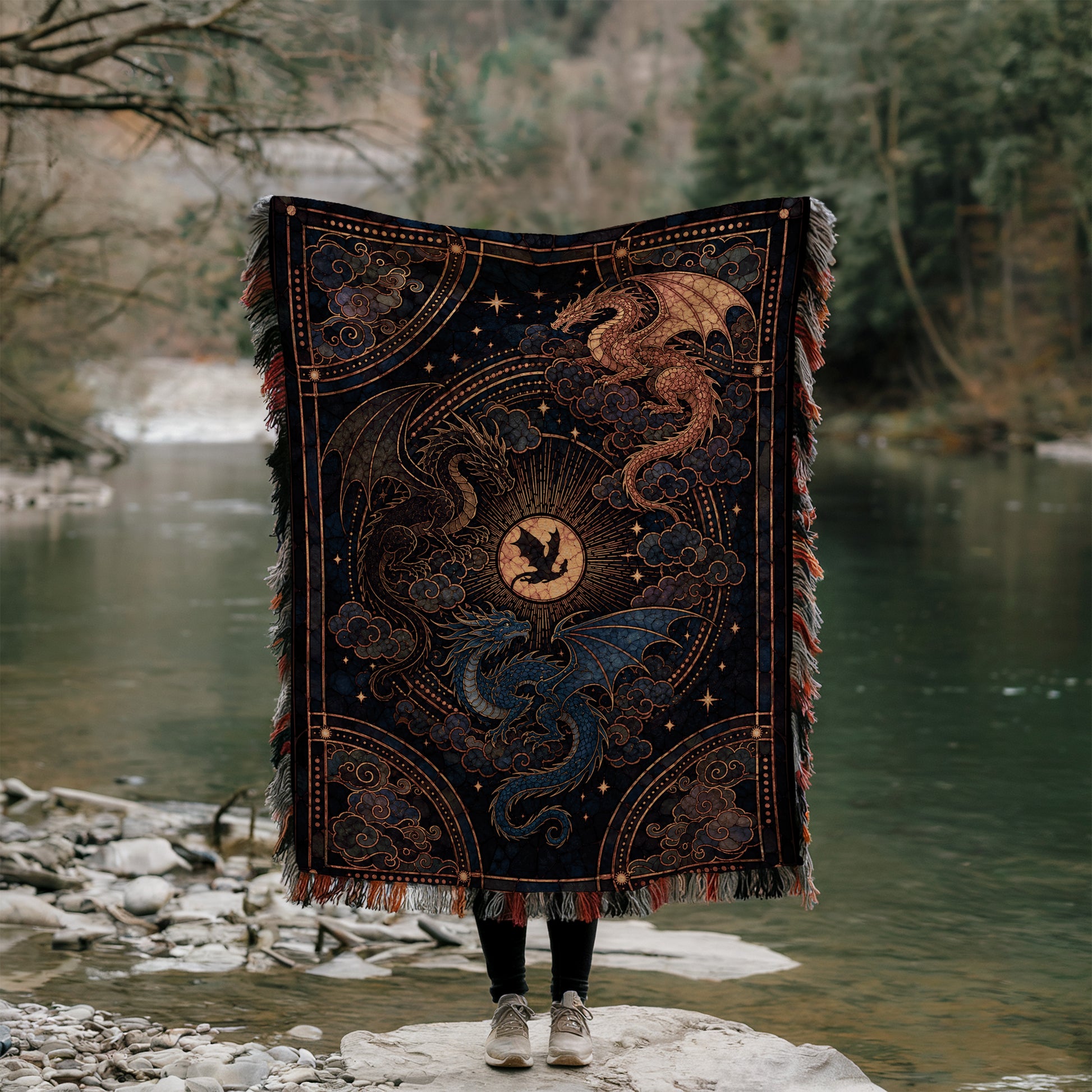 A person stands on a rock, holding a large, intricately designed blanket featuring a dragon design. The blanket is adorned with a central sun motif surrounded by swirling patterns and a dragon-like creature. The person is positioned in front of a body of water, with trees and foliage visible in the background.