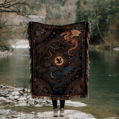 A person stands on a rock, holding a large, intricately designed blanket featuring a dragon design. The blanket is adorned with a central sun motif surrounded by swirling patterns and a dragon-like creature. The person is positioned in front of a body of water, with trees and foliage visible in the background.