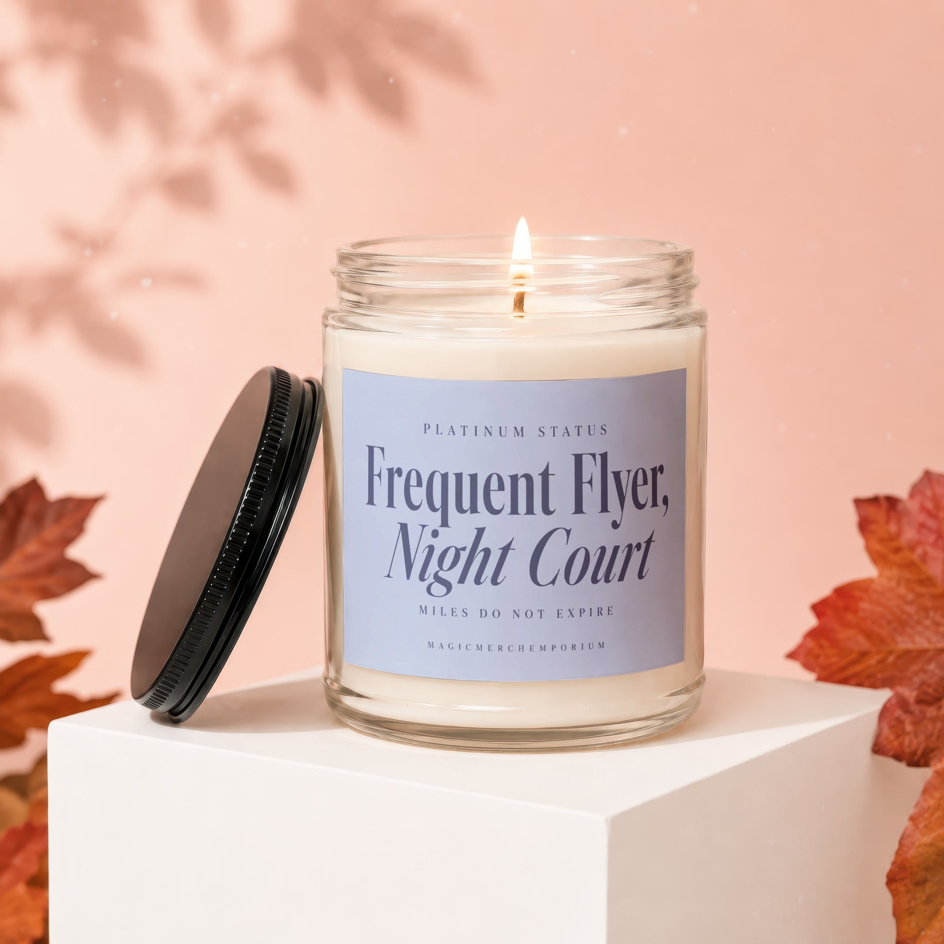A lit jar candle with a purple label rests on a white pedestal, surrounded by autumn leaves.