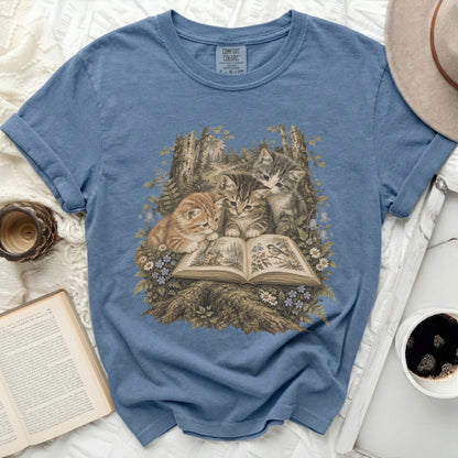 Three tabby kittens read a book in a forest clearing with flowers.