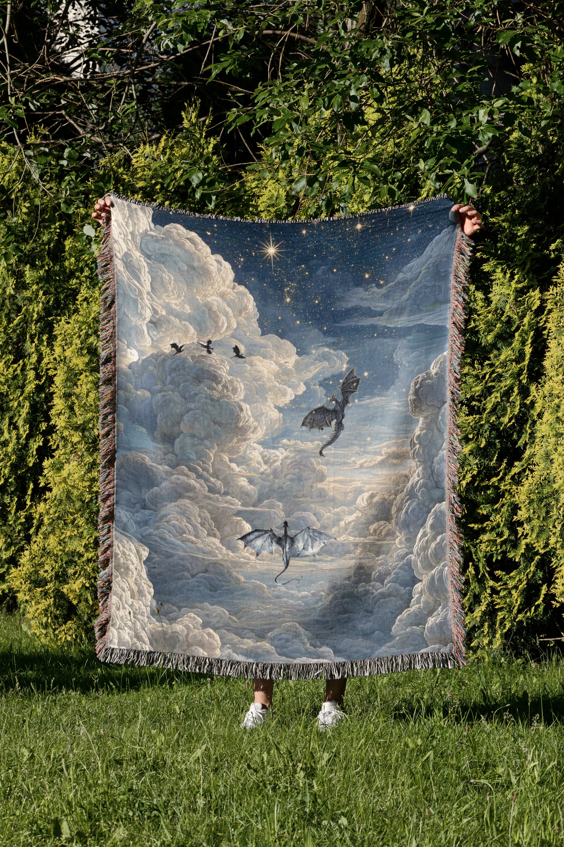 A person is holding up a large, colorful blanket with a dragon and clouds design, standing in a grassy area surrounded by trees.