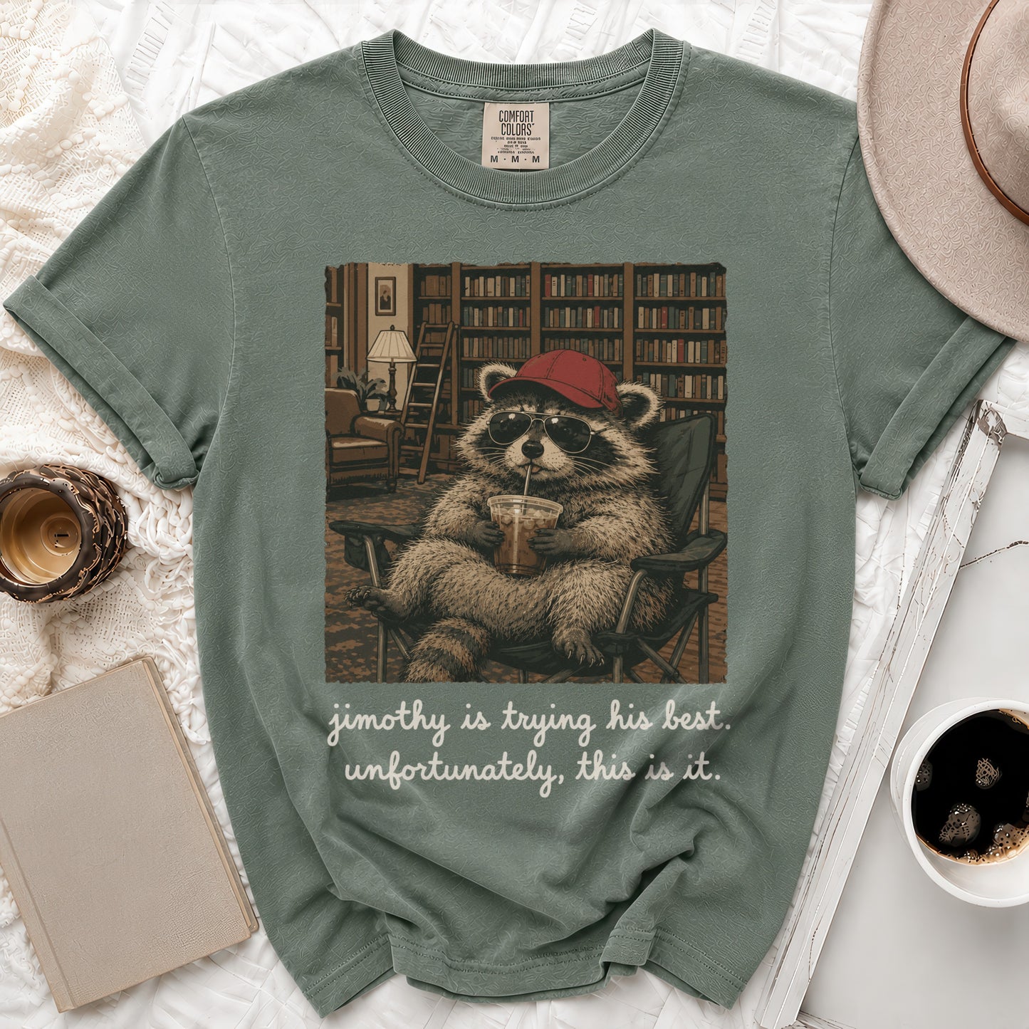 T-shirt graphic of a raccoon wearing a red hat and sunglasses drinking from a cup.