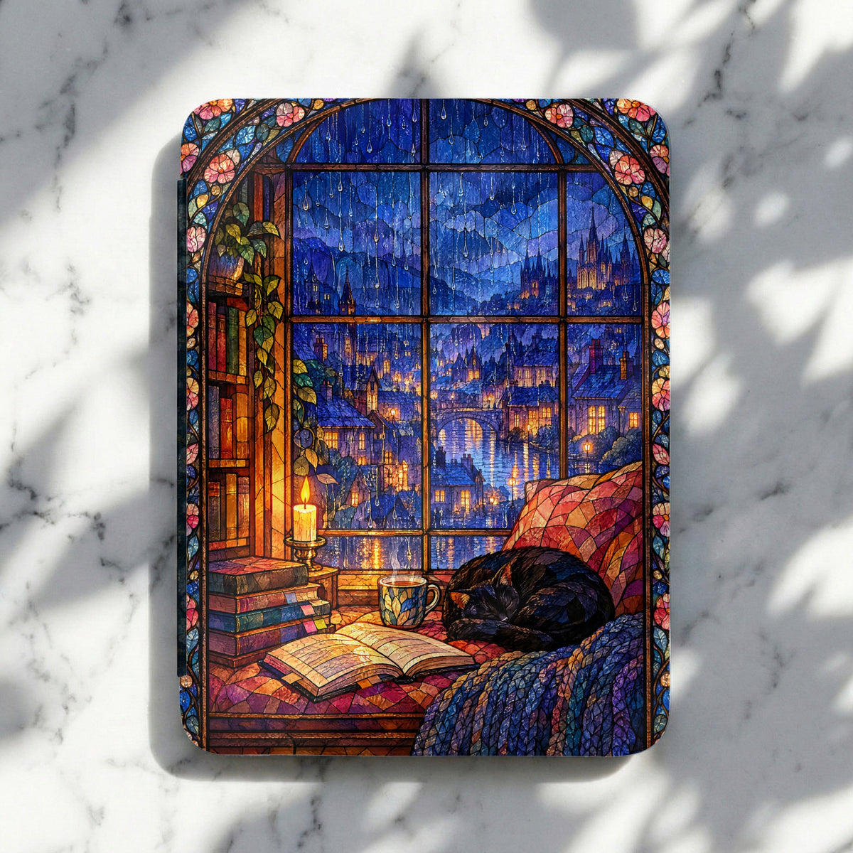 A Kindle case features a colorful stained glass illustration of a sleeping cat, stacked books, and a rainy medieval city view.