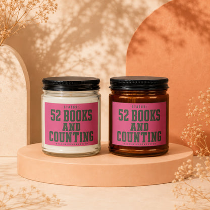 Two candles, one clear and one amber, with pink labels reading "Status: 52 Books And Counting."