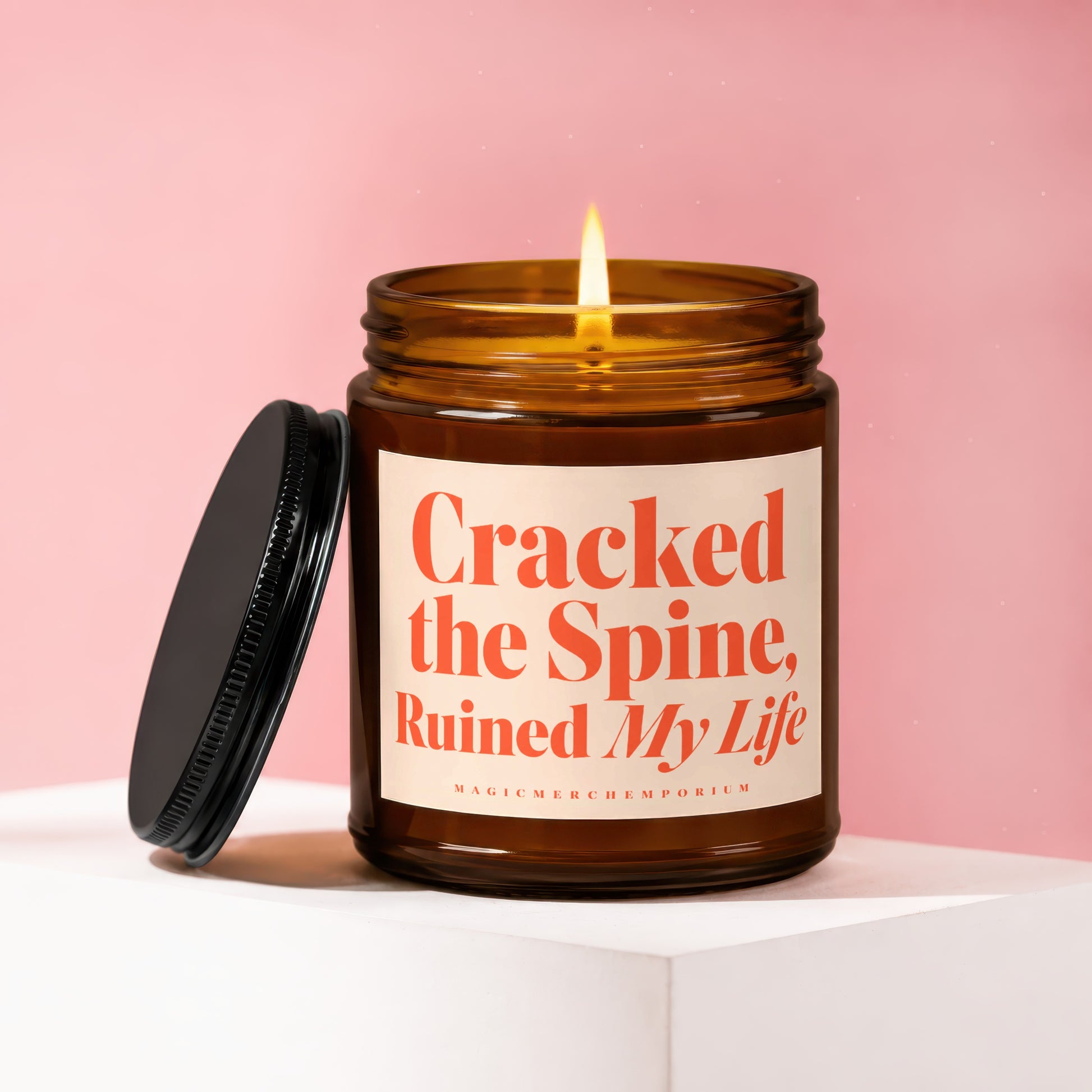 Brown glass candle with lit flame and black lid, labeled "Cracked the Spine, Ruined My Life."