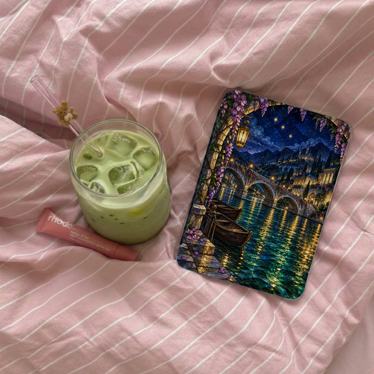Glass of green drink with ice, lip balm, and art print on pink striped fabric.
